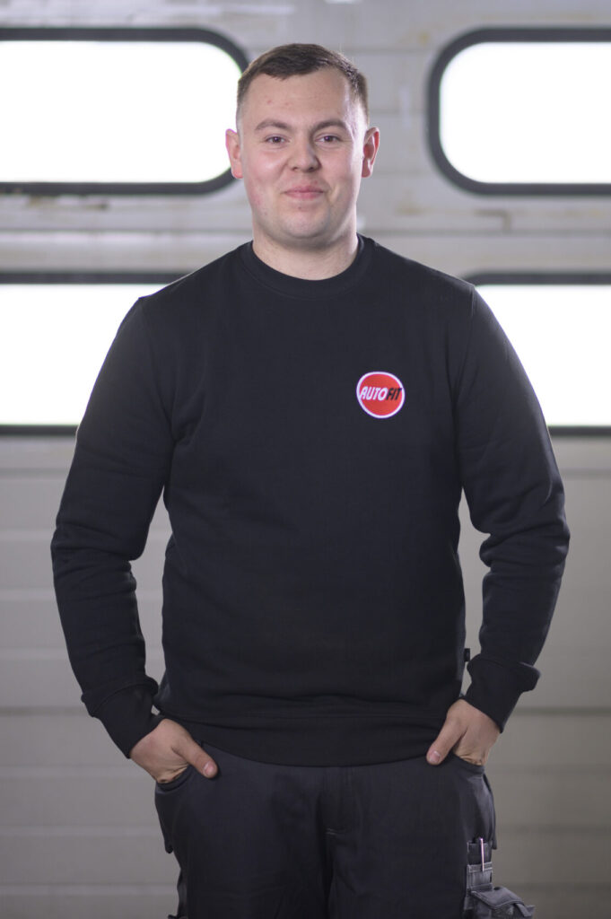 Eine Person steht in schwarzer Arbeitskleidung mit AUTO FIT-Logo vor einem Garagentor und hat beide Hände in den Hosentaschen.