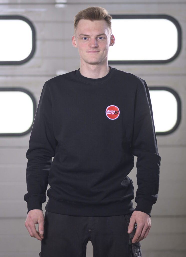 Eine Person steht in schwarzer Arbeitskleidung mit AUTO FIT-Logo vor einem Garagentor.