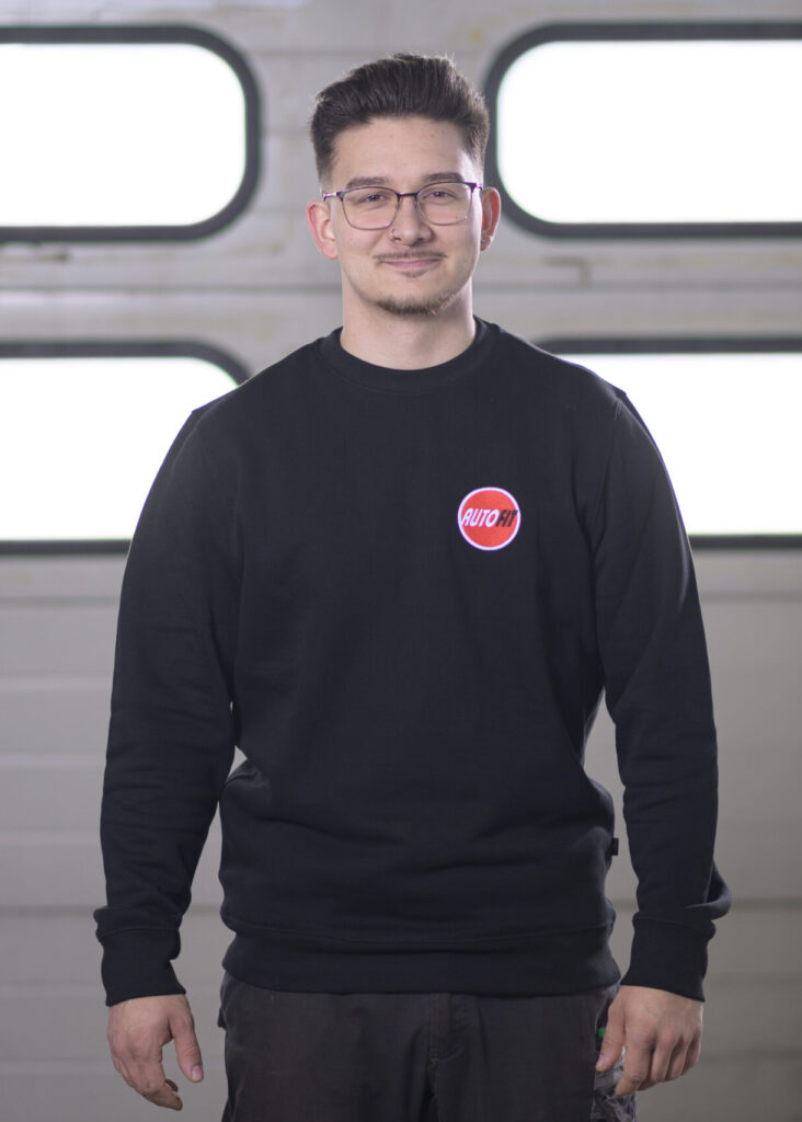 Eine Person steht in schwarzer Arbeitskleidung mit AUTO FIT-Logo vor einem Garagentor.