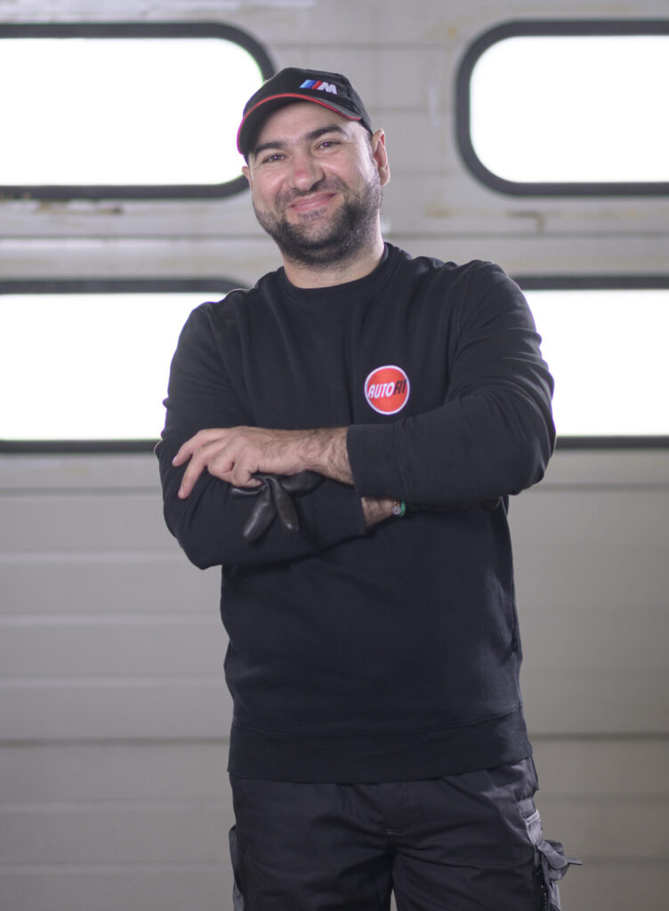 Eine Person in schwarzer Arbeitskleidung mit AUTO FIT-Logo und Mütze steht mit verschränkten Armen und hält Arbeitshandschuhe vor einem Garagentor.