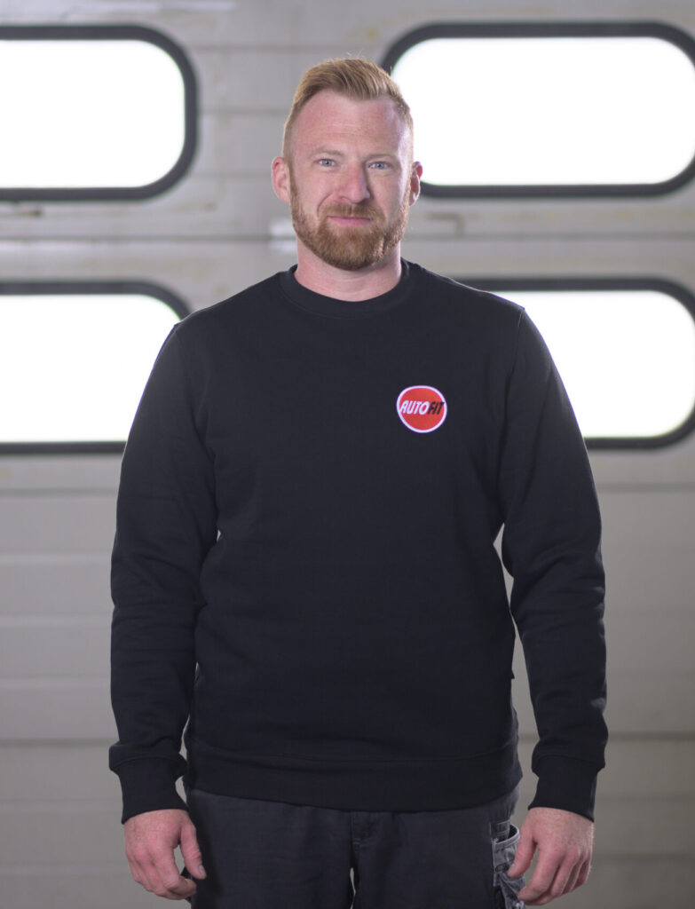 Eine Person steht in schwarzer Arbeitskleidung mit AUTO FIT-Logo vor einem Garagentor.