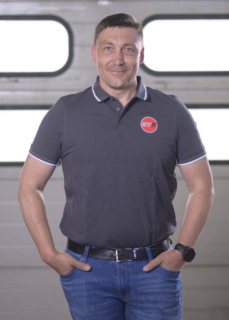 Eine Person steht vor einem Garagentor, trägt ein graues Poloshirt mit AUTO FIT-Logo, eine Jeans und hat die Hände in den Hosentaschen.