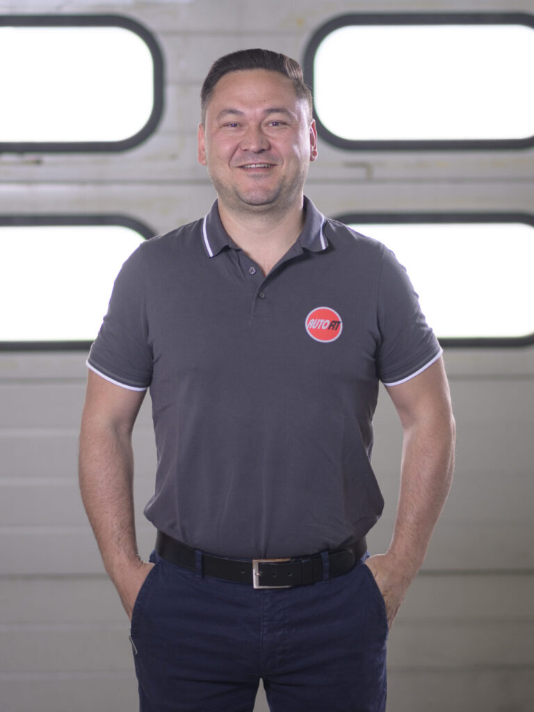 Eine Person steht vor einem Garagentor, trägt ein graues Poloshirt mit AUTO FIT-Logo und hat die Hände in den Hosentaschen.
